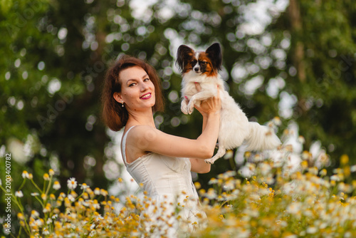A woman in a white dress holds a Papillon dog in her arms in a field of chamomile.