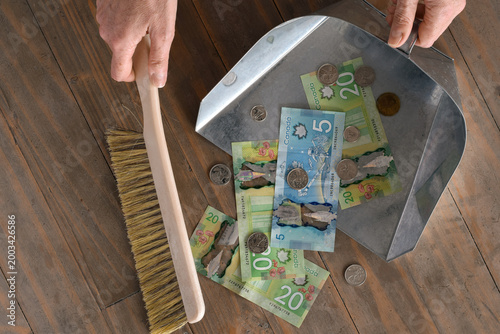 Hands sweeping Canadian dollar bills from floor into dustpan, symbolizing financial loss, rising expenses, debt, or cost of living pressure. Concept image for inflation, recession, taxes, or money man