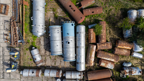 Top down aerial view of industrial storage yard featuring abandoned cylindrical metal tanks and pressure vessels in various states of rust and silver finish on concrete and grass surface.