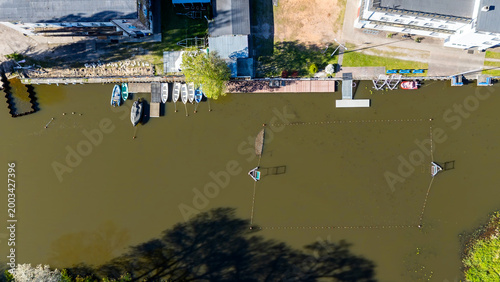 Billede på lærred Aerial top-down flat lay view of a river marina with colorful rowing boats and motorboats moored at a wooden pier, waterfront buildings, green trees, and long shadows on murky water