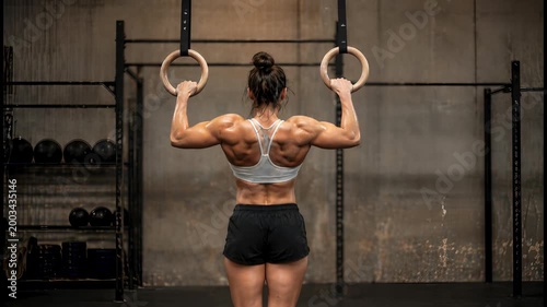 Strong woman performing gymnastic ring pull-ups for back muscle workout in gym