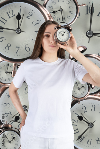 Woman holding an alarm clock looking into camera, surrounded by many clock faces