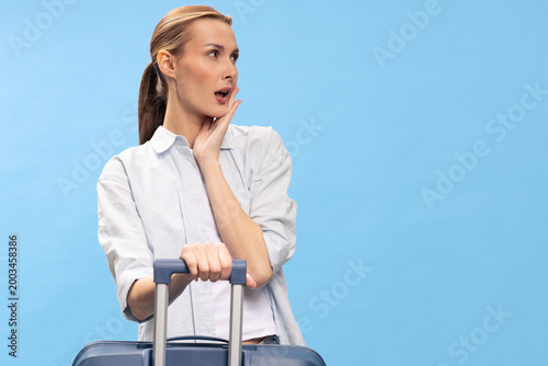 Obraz na plátne Surprised woman in white shirt holding suitcase handle looks aside with open mouth on blue background