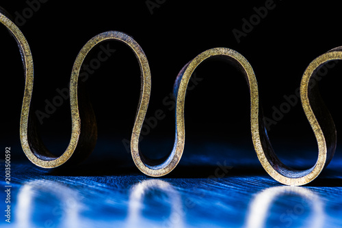 Closeup of a ribbon curled brass flat bar with noble patina, placed on a metal surface in industrial lighting, forming abstract curved shapes