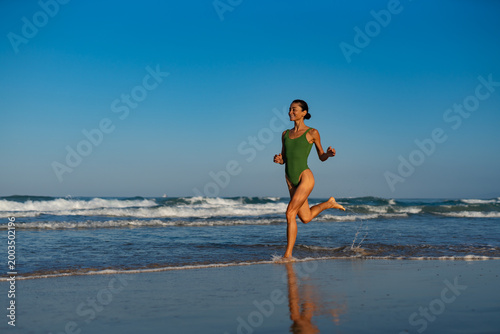 Cheerful woman in green swimsuit running along the beach in shallow water, enjoying the sunlight and ocean waves on a bright summer morning, smiling with freedom and energy.