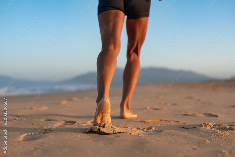 Fototapeta premium Close-up of fit woman’s legs walking barefoot on beach at sunrise, sand sticking to her foot, with soft golden light and mountains blurred in the background, early morning fitness concept.