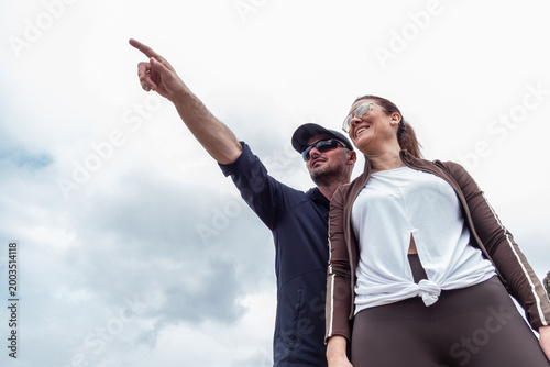 Fotografi Man and woman outdoor pointing finger up against a cloudy sky, looking ahead wit