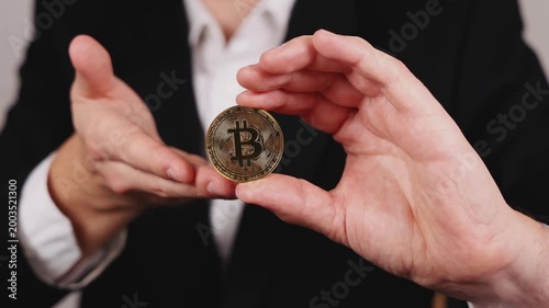 Une main tient une pièce de Bitcoin dorée entre deux doigts. En arrière plan, une personne en costume et chemise blanche.