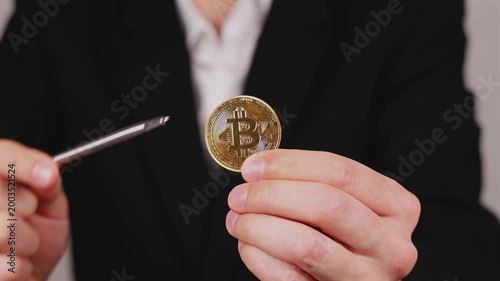 Une main montre une pièce de Bitcoin dorée avec un stylo. En arrière plan, une personne en costume et chemise blanche. 