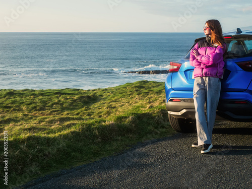 Young woman in colorful jacket is standing by a blue rented car and looking at stunning nature scenery with rough ocean coast at sunset. Mullaghmore, county Sligo. Travel and tourism in Ireland.