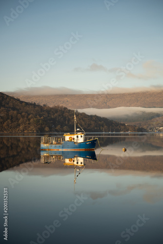 Slika na platnu Fishing boat reflected on calm waters of a Scottish loch, with mountains and soft morning light