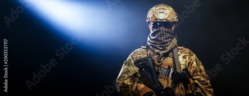 Fotografía Soldier in full gear holding assault rifle on dark smoke background