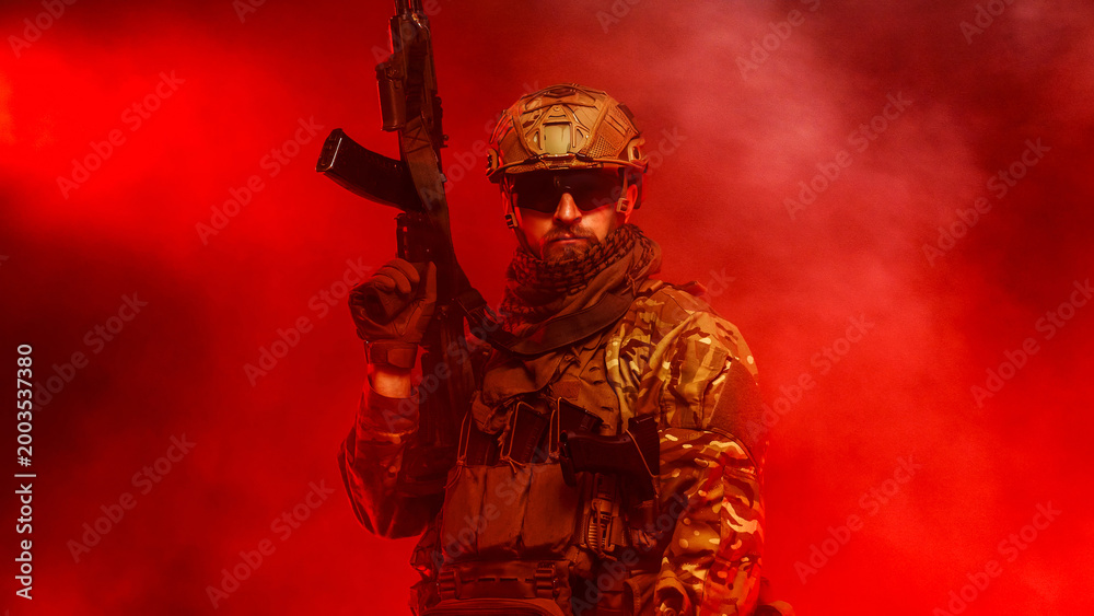 Fototapeta premium Military soldier with assault rifle standing on red smoky background. Concept defense, tension and security forces. 