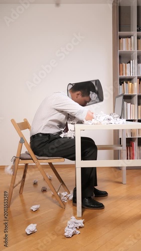 Vertical video businessman feeling overwhelmed and frustrated, sitting at his desk with a wastepaper basket full of crumpled paper falling over his head, representing work stress and failure