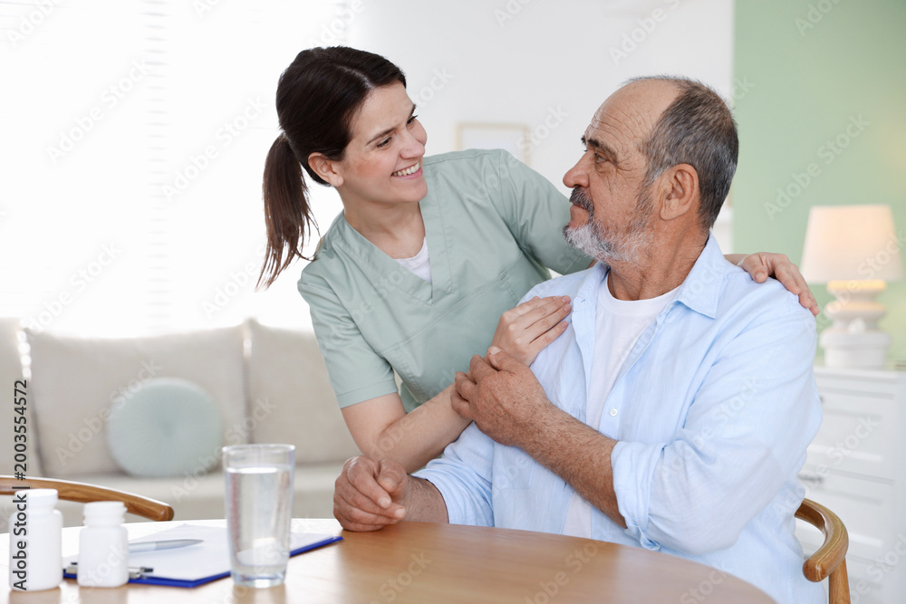 Obraz premium Smiling care worker supporting senior man at wooden table at nursing home