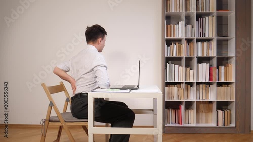 Man feeling lower back pain while sitting at a desk working on a laptop, highlighting the importance of ergonomics and proper posture for health and well being in an office environment