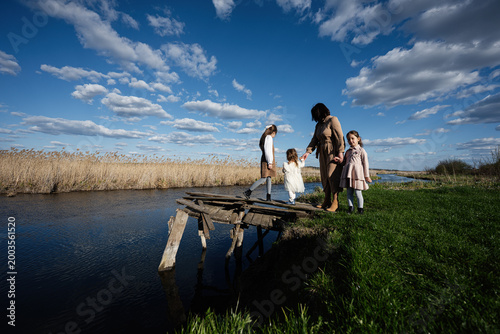 Εκτύπωση καμβά Mother and daughters walking on a rickety wooden bridge by a river in a rural la