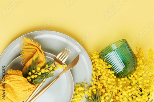 Bright spring table setting with mimosa flowers ceramic plates golden cutlery yellow napkin and green glass festive seasonal composition