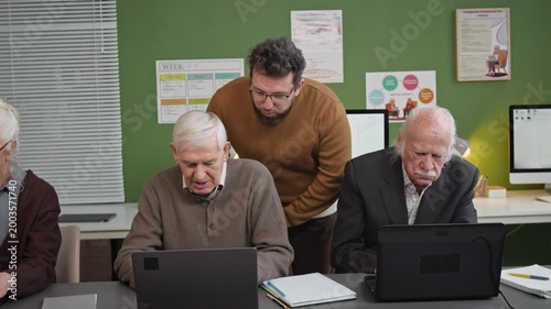 Following front view shot of caring male teacher overseeing group of seniors using laptops during computer literacy class and assisting adult students one by one