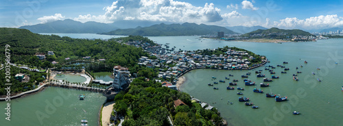 An aerial view of a coastal village in Hon Mieu, Nha Trang, Vietnam, with numerous fishing boats docked in the turquoise bay. The village is nestled between lush green hills and the sea.