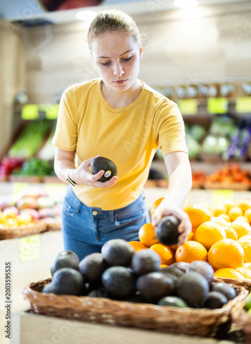 Картината върху платно Young woman comes to the farm store with fruits and vegetables and carefully chooses a ripe avocado on the recommendation of a nutritionist