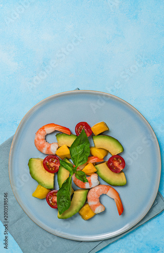 Fresh salad with shrimps, avocado, mango, and cherry tomatoes on a blue plate, blue background with copy space, top view.