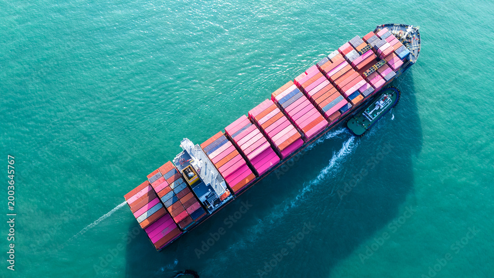 Fototapeta premium Overhead aerial view of a massive cargo ship fully loaded with pink and red containers, assisted by a tugboat in the turquoise ocean. Global maritime logistics and supply chain concept.
