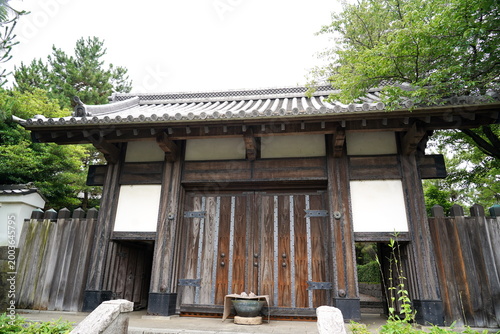 Photos 歴史を感じさせる重厚な日本の伝統的な木造の門。美しい瓦屋根と白い漆喰壁が特徴的な建築物で周囲には夏を感じさせる青々とした樹木が広がっている。