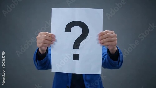A person in a blue jacket holds a white paper with a large question mark in front of their face, concealing their identity