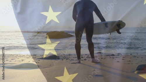Male surfer walking to shoreline adjusting fins for sports with flag overlaying scene yellow stars