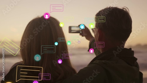 Man raising phone capturing sunset with woman for social, causing avatars and chat bubbles drifting