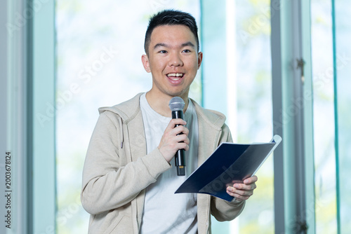 An Asian young man smiles broadly while speaking into a microphone and holding a blue folder in a university lecture hall, perfect for student presentation, public speaking, or campus event concepts.