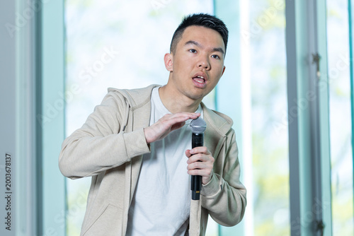 An Asian young man taps a handheld microphone to test it in a university lecture hall, perfect for event setup, audio equipment, or campus event preparation concepts.