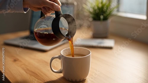 A person is pouring hot beverage from a glass carafe into a white ceramic mug on a wooden desk near a window