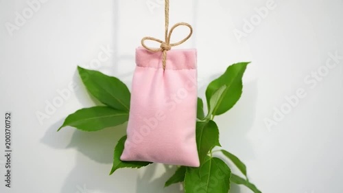 A small, pink fabric pouch hangs from a natural twine string, adorned with green leaves, against a white background