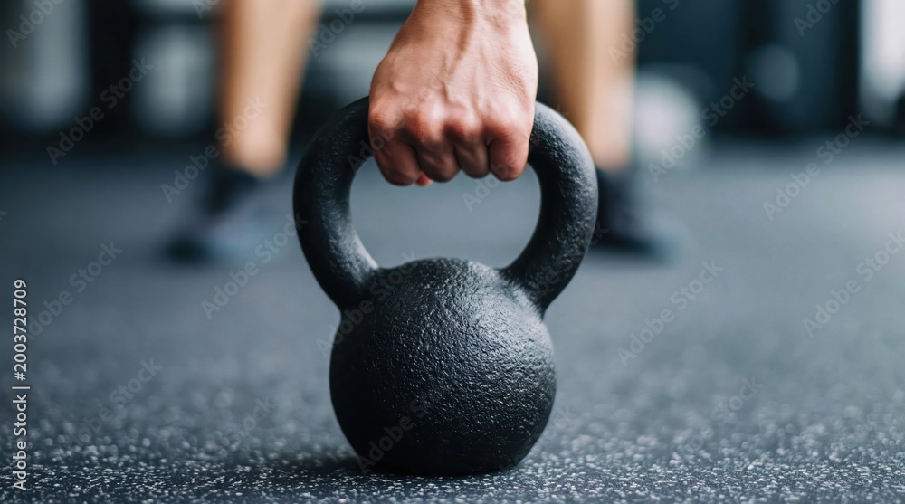 Naklejka premium Exercising hand gripping a heavy black kettlebell on a gym floor, concept of fitness, strength, and training