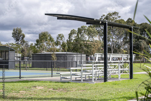 Konstfotografi Tiered metal spectator bleachers under a modern  cantilevered shade at a suburban sports park, overlooking a fenced outdoor court, a grey water tank