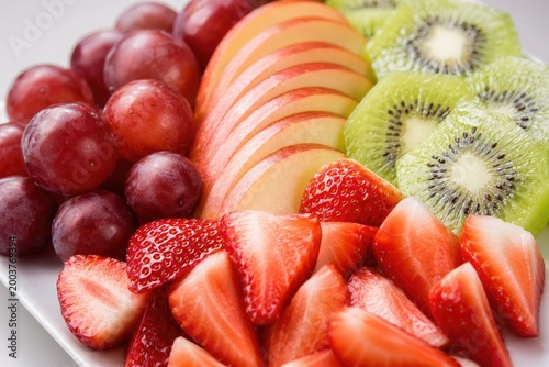 Freshly sliced vibrant fruit platter featuring red grapes, sliced strawberries, kiwi halves, and apple slices arranged neatly on white plate, healthy snacking concept, bright studio lighting