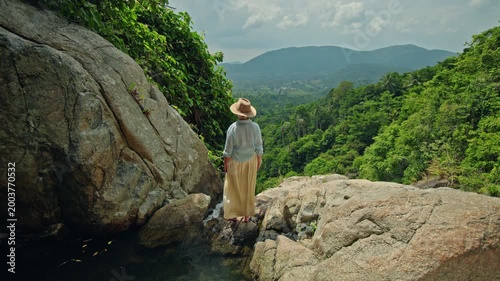 Middle-aged woman standing barefoot on rocky ledge near tropical waterfall overlooking lush green valley and distant mountains under bright cloudy sky