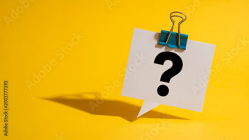 A question mark on a white speech bubble paper is held by a blue clip against a bright yellow background