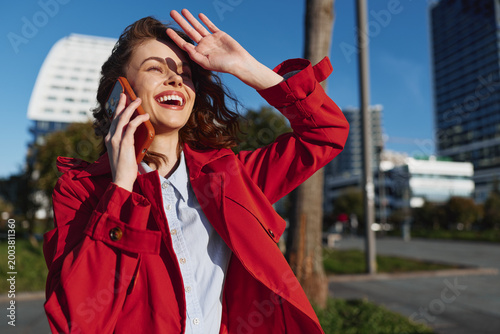 Canvastavla Smiling woman in a red jacket talks on a mobile phone outdoors, urban park setting, candid lifestyle and fashion moment, sunny atmosphere, bright cheer