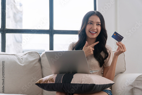 Happy Asian woman sitting on sofa with laptop pointing at credit card for online shopping. Beautiful young female smiling and recommending electronic payment transaction at home.