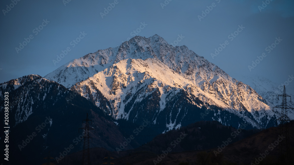 Obraz premium Majestic Winter Alpine Landscape at Sunset with Pine Trees, Sun-Kissed Snow Ridges Above Dark Mountain Woods