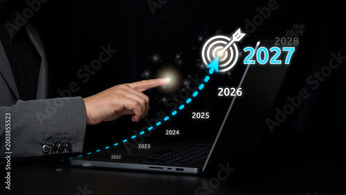 Hand points at a glowing 2027 target icon above laptop, combining business growth timeline with financial analytics and future performance tracking.