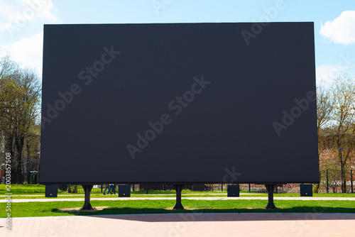 A large digital screen is positioned in a park area surrounded by grass and trees. The screen remains blank against a clear blue sky, suggesting a potential for upcoming events or advertisements