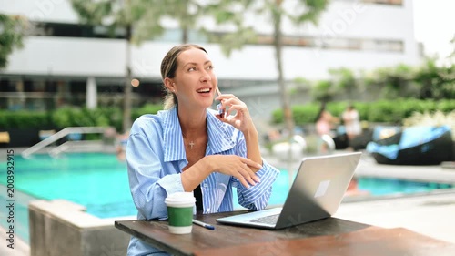Business woman working remotely on laptop by pool, talking on phone. Digital nomad lifestyle, online work, productivity, travel, luxury resort