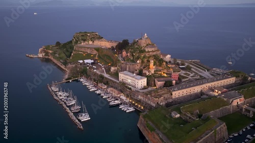 Aerial view of the Old Fortress in Corfu Town at twilight, featuring the historic Venetian citadel, illuminated architecture, and docked sailboats in the marina on the Ionian Sea