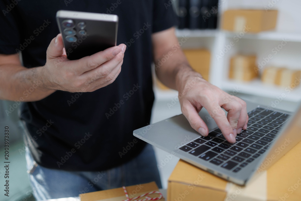 Fototapeta premium Man packs boxes at desk with laptop nearby. Focused, he arranges parcels with care and efficiency. Bright room features shelves, supplies, and organized chaos. Ideal for e-commerce, logistics