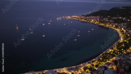 Twilight aerial drone shot over Garitsa Bay in Corfu, Greece. Sailboats anchored in calm waters next to the illuminated glowing coastal city streets and coastline