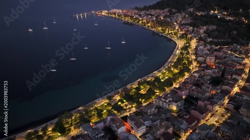 Aerial night view of Garitsa Bay in Corfu Town, Greece. Illuminated city streets, coastal promenade, and sailboats anchored in the dark Ionian Sea at twilight